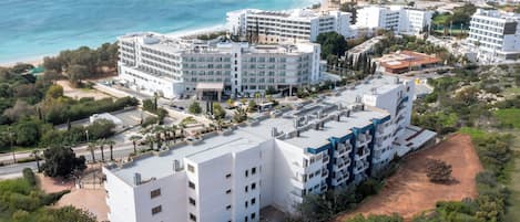 A coastal hotel with multiple white buildings, a tennis court, and a beach in the background.