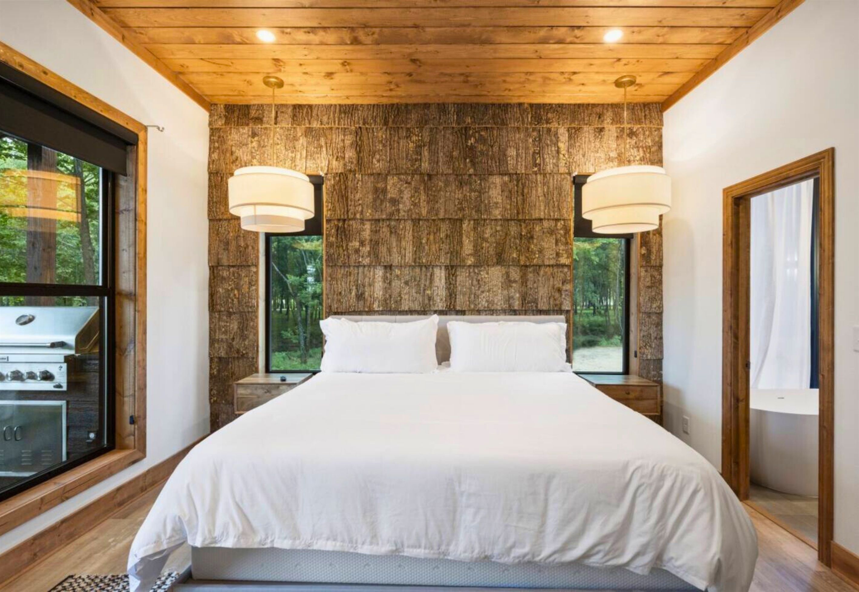 A bedroom with a large bed, two bedside lamps, a view of the outdoors through a window, and a wooden ceiling.