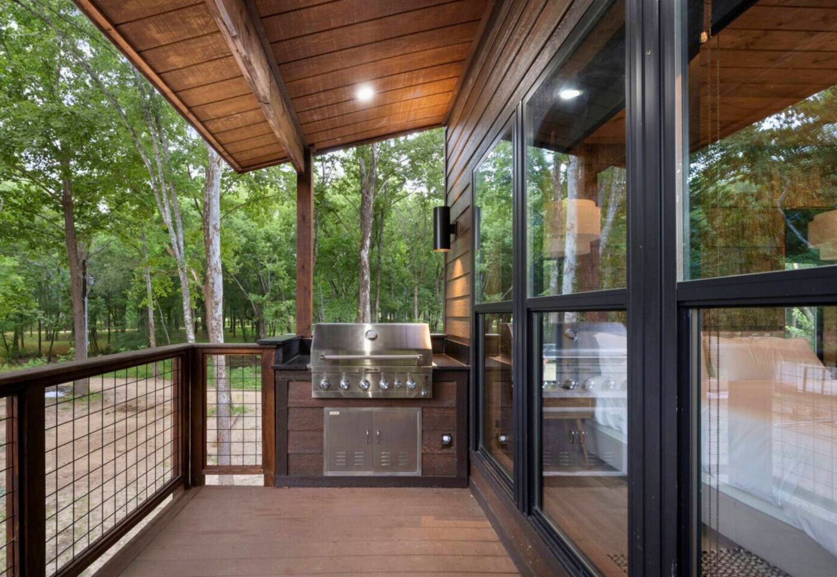 An outdoor deck with a grill and a glass enclosure, surrounded by trees.