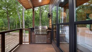 Terraza exterior con una parrilla y cerramiento de vidrio, rodeada de árboles.