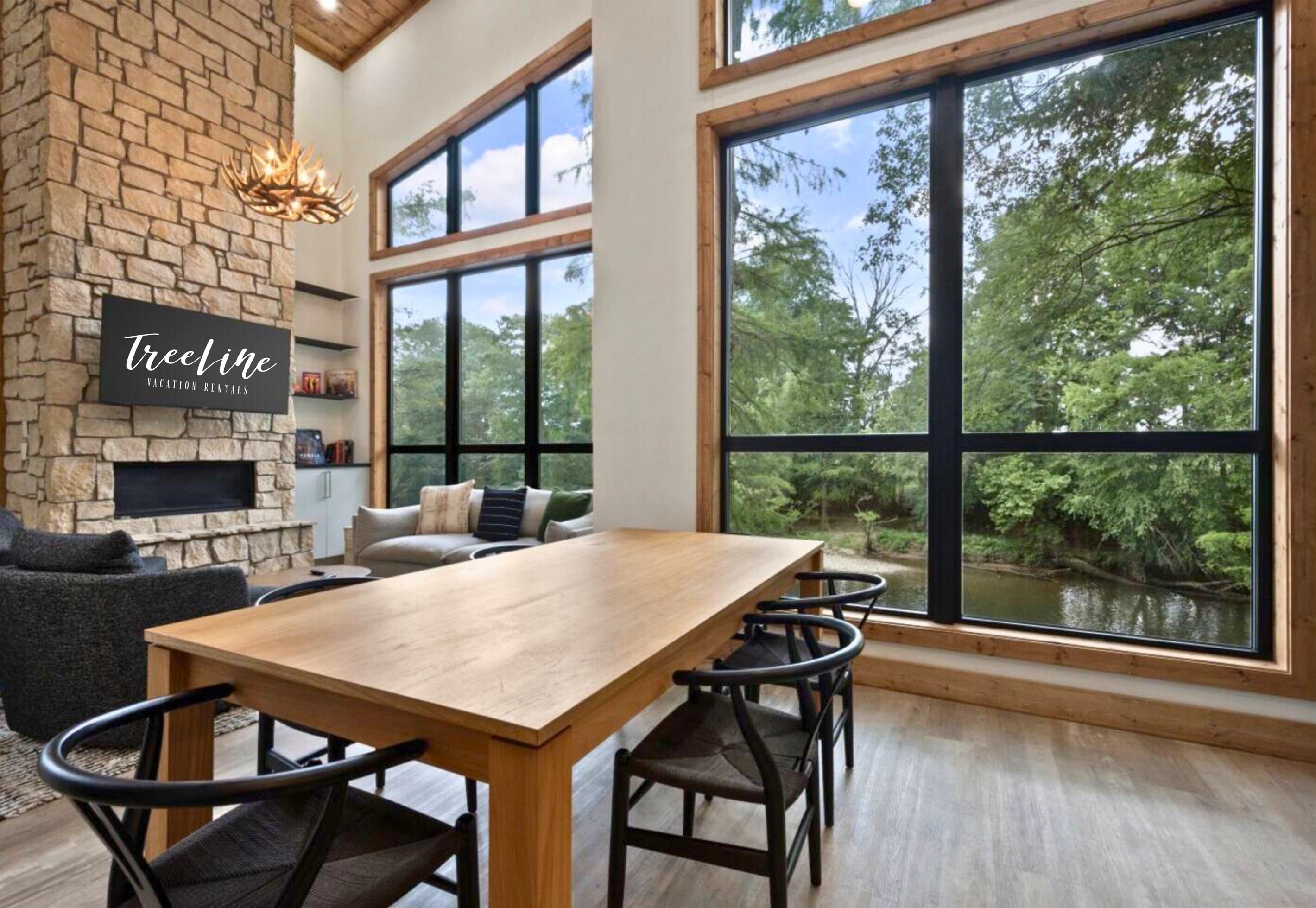 A spacious room with a large wooden table, comfortable seating, a stone fireplace, and large windows offering a view of the outdoors.