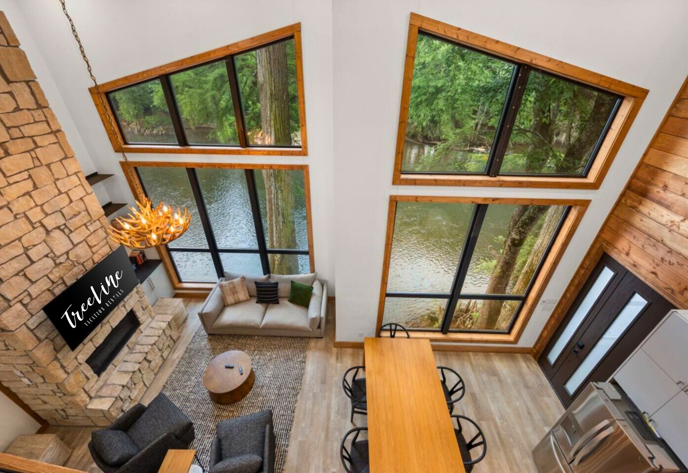 A modern interior with a stone fireplace, a sofa, a dining table, and large windows offering a view of a forested area.
