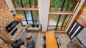 Un interior moderno con chimenea de piedra, un sofá, una mesa de comedor y ventanas amplias con vistas a un área boscosa.