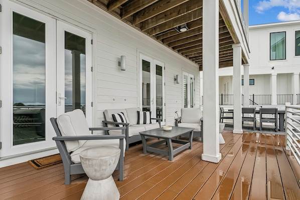 A covered outdoor area with white walls, wooden flooring, and furniture including a sofa, chairs, and a table.