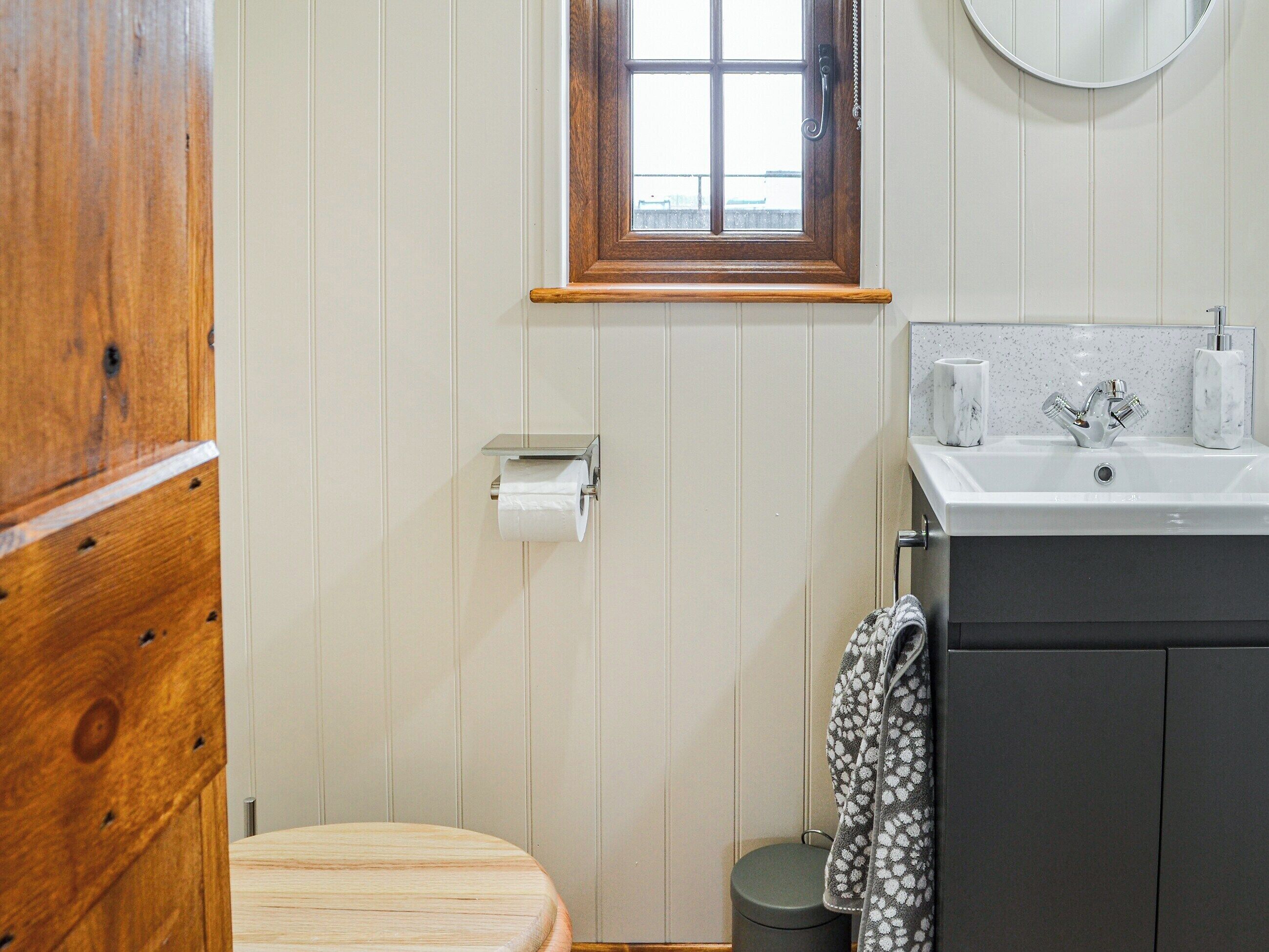 Un baño con inodoro de madera, un dispensador de papel higiénico blanco, un lavamanos con encimera de mármol y un espejo sobre el lavamanos.