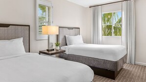 Ein Hotelzimmer mit zwei Betten, einem Nachttisch mit Lampe, einem Fenster mit Vorhängen und Blick auf Palmen.