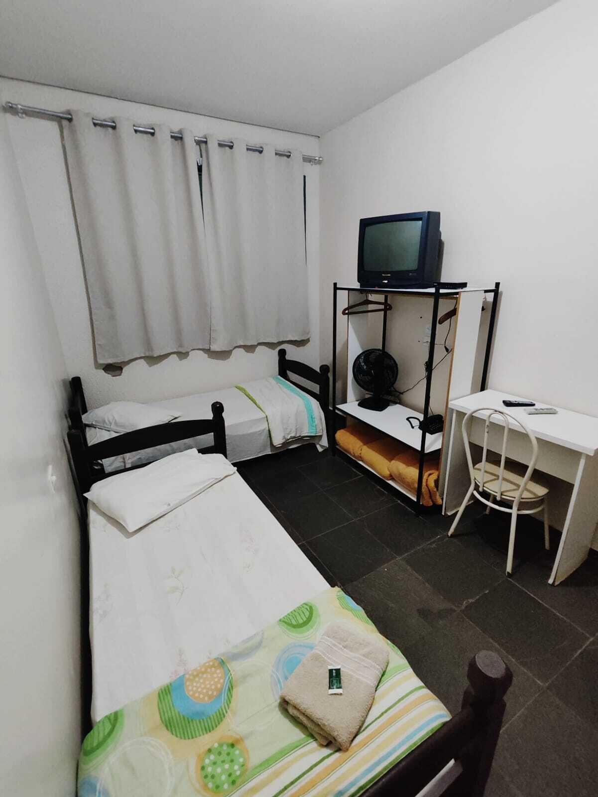 Una habitación pequeña y limpia con dos camas individuales, un escritorio, una silla, un televisor y un ventilador.