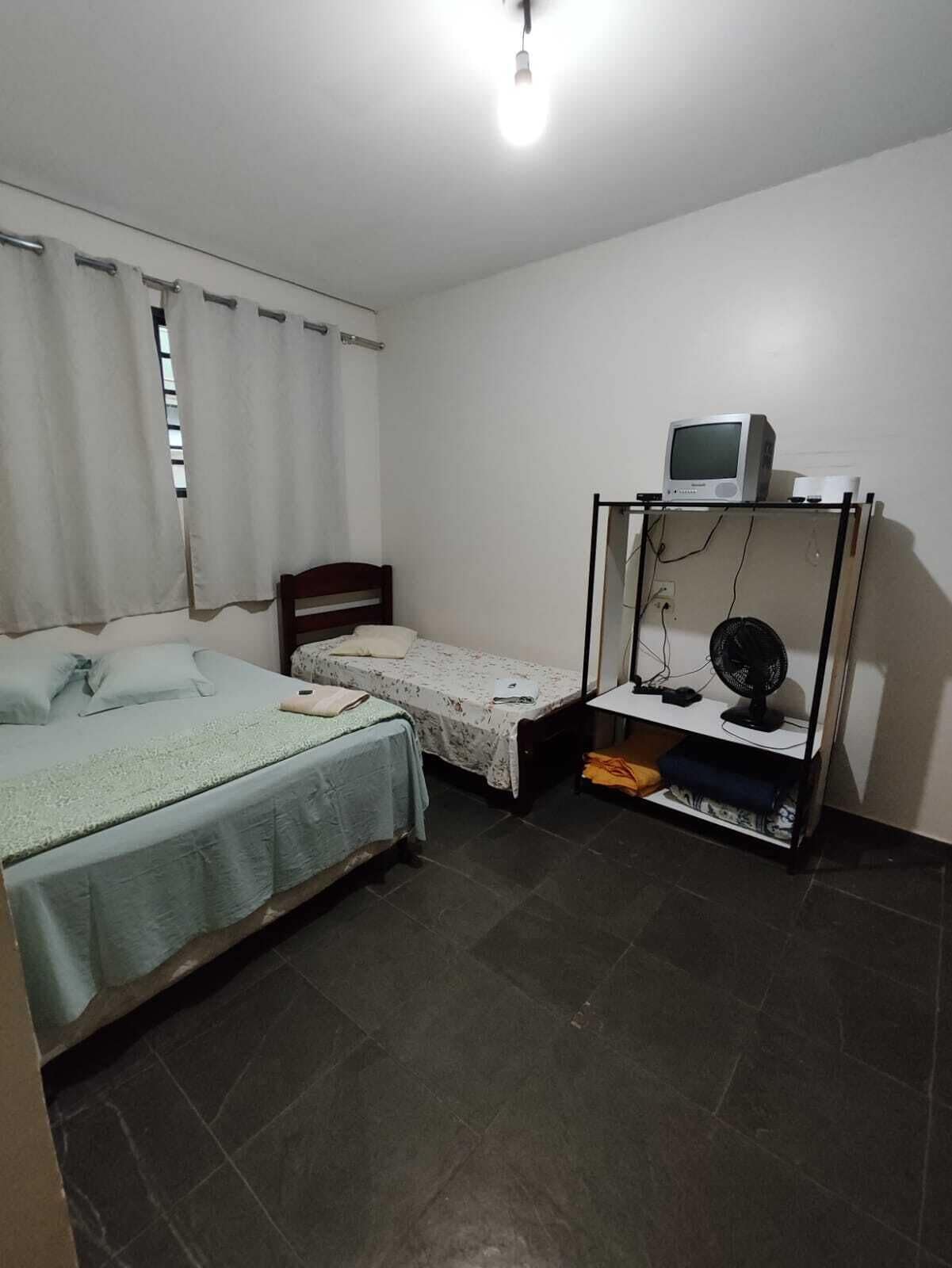 A small, simple room with two beds, a TV on a stand, a fan, and a window with curtains.