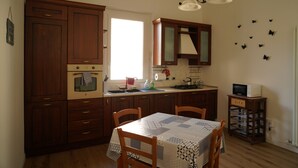 Una cocina con armarios de madera, una mesa de comedor con sillas, un microondas sobre un carrito y una ventana con luz natural.