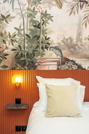 Quarto de hotel com cama, luminária na parede e um mural de um pássaro e folhagem.