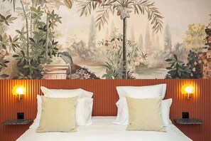 Un dormitorio con un mural de paisaje tropical, dos mesitas de noche con lámparas y una cama con ropa de cama blanca y almohadas color beige.