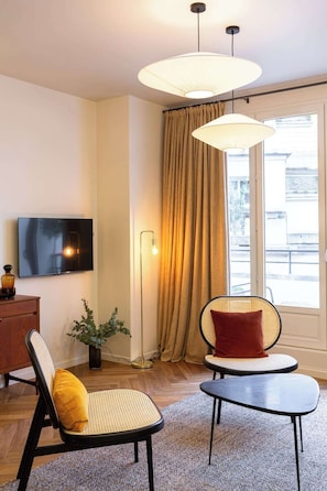 Una sala de estar moderna con un televisor de pantalla plana, una lámpara de pie grande, un sofá con un cojín amarillo, una mesa redonda y una ventana con cortinas.