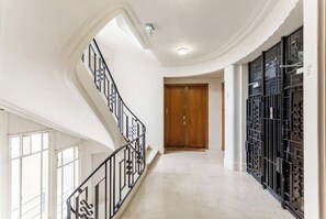 Un pasillo amplio con una escalera curva, una puerta de madera y un muro con contenedores metálicos.