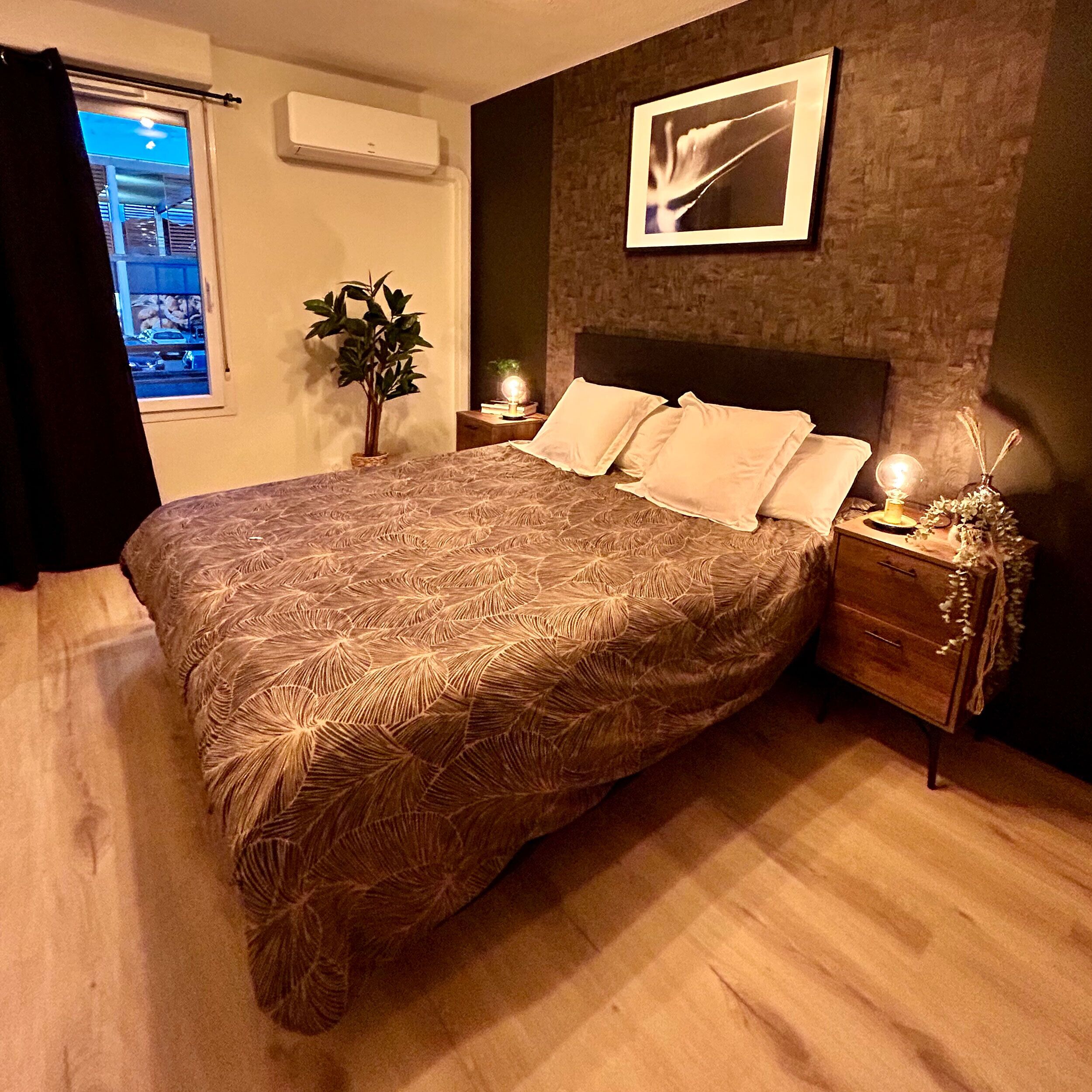 A bedroom with a large bed, a bedside table with a lamp, a wall-mounted air conditioner, a framed picture, and a window with a view of the outside.