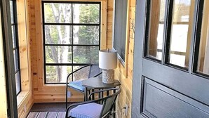 A wooden cabin interior with a small table and chair, a window with a view of trees, and a door.