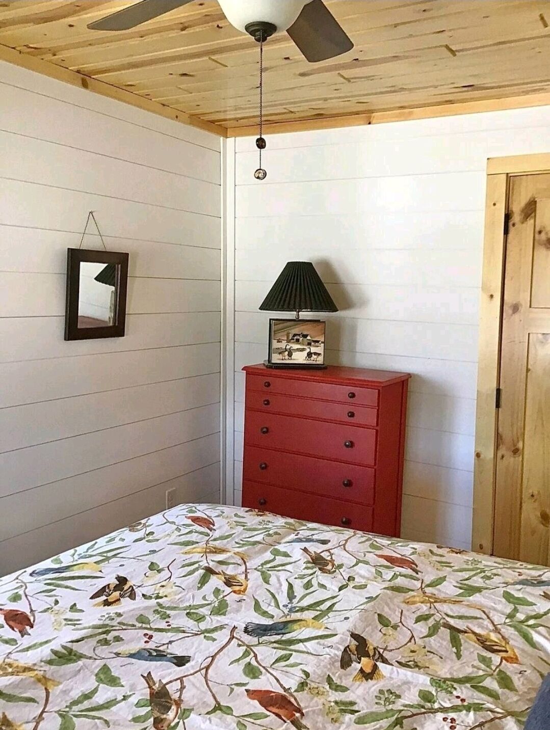 A bedroom with a wooden ceiling, a bed with a floral bedspread, a red dresser, a wall-mounted mirror, and a ceiling fan.