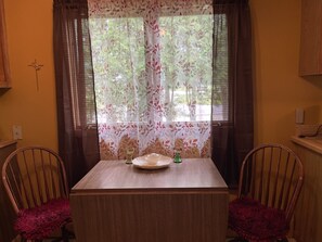 Et spiseområde med et bord og to stole, et vindue med blomstermønstrede gardiner og en lampe monteret på væggen.