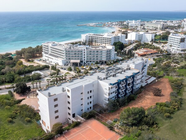 Ein Hotel an der Küste mit mehreren weißen Gebäuden, einem Tennisplatz und einem Strand im Hintergrund.