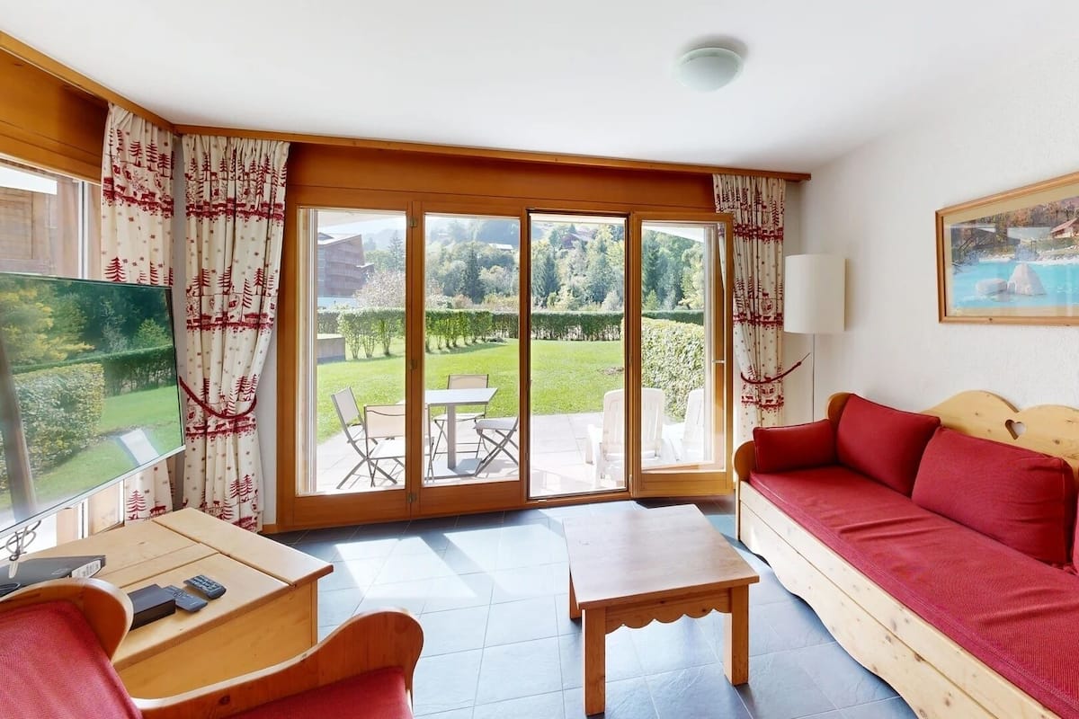 A room with a wooden sofa, a small table, a TV, and a view of a garden through large sliding doors.