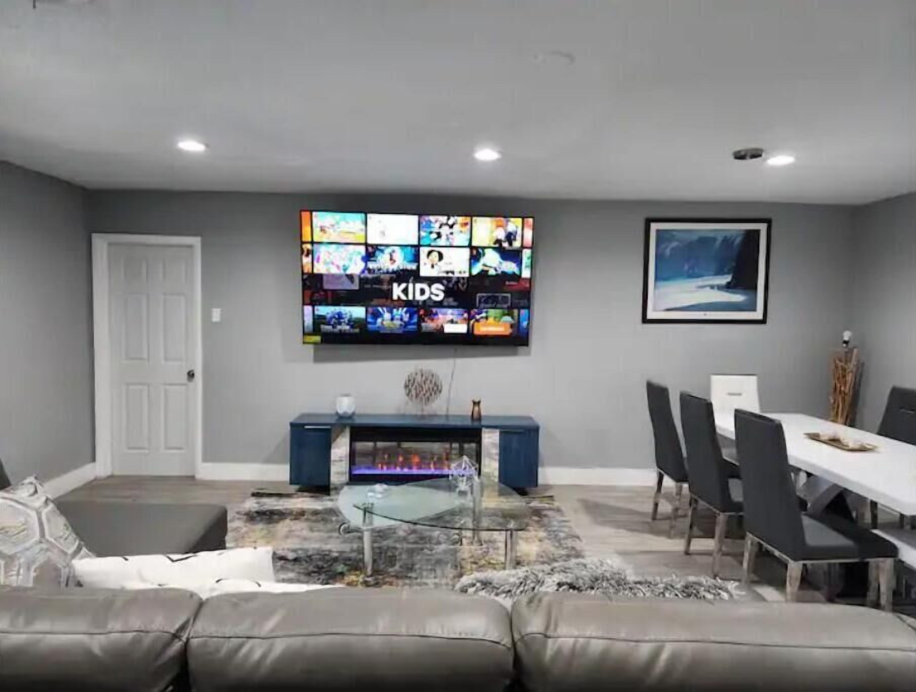 A living room with a large TV displaying children's channels, a dining area with a table and chairs, and a grey couch.