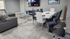 A modern living room with a grey sofa, a glass coffee table, a dining table with chairs, a flat-screen TV mounted on the wall, and a framed picture above the TV. - The Perfect Vacation Getaway ! (Miami)