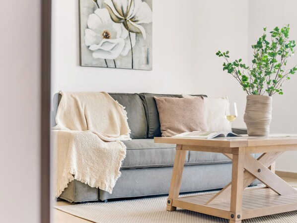 A living room with a grey sofa, a wooden coffee table, a white vase with green plants, and a framed floral artwork on the wall.
