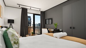 A modern hotel room with a large bed, a desk, a chair, a TV, and a balcony with a view of buildings. - Cozy Studio in New Development - Backup Generator (Cape Town)