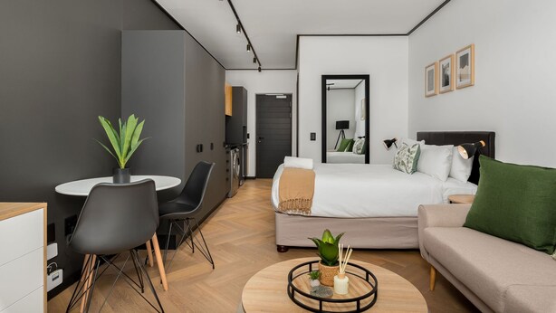 A modern hotel room with a bed, a sofa, a dining table with chairs, a small kitchen area, and a mirror. - Cozy Studio in New Development - Backup Generator (Cape Town)