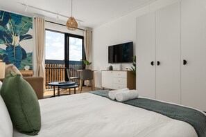 A modern hotel room with a large bed, a desk, a chair, a TV, and a balcony with a view.