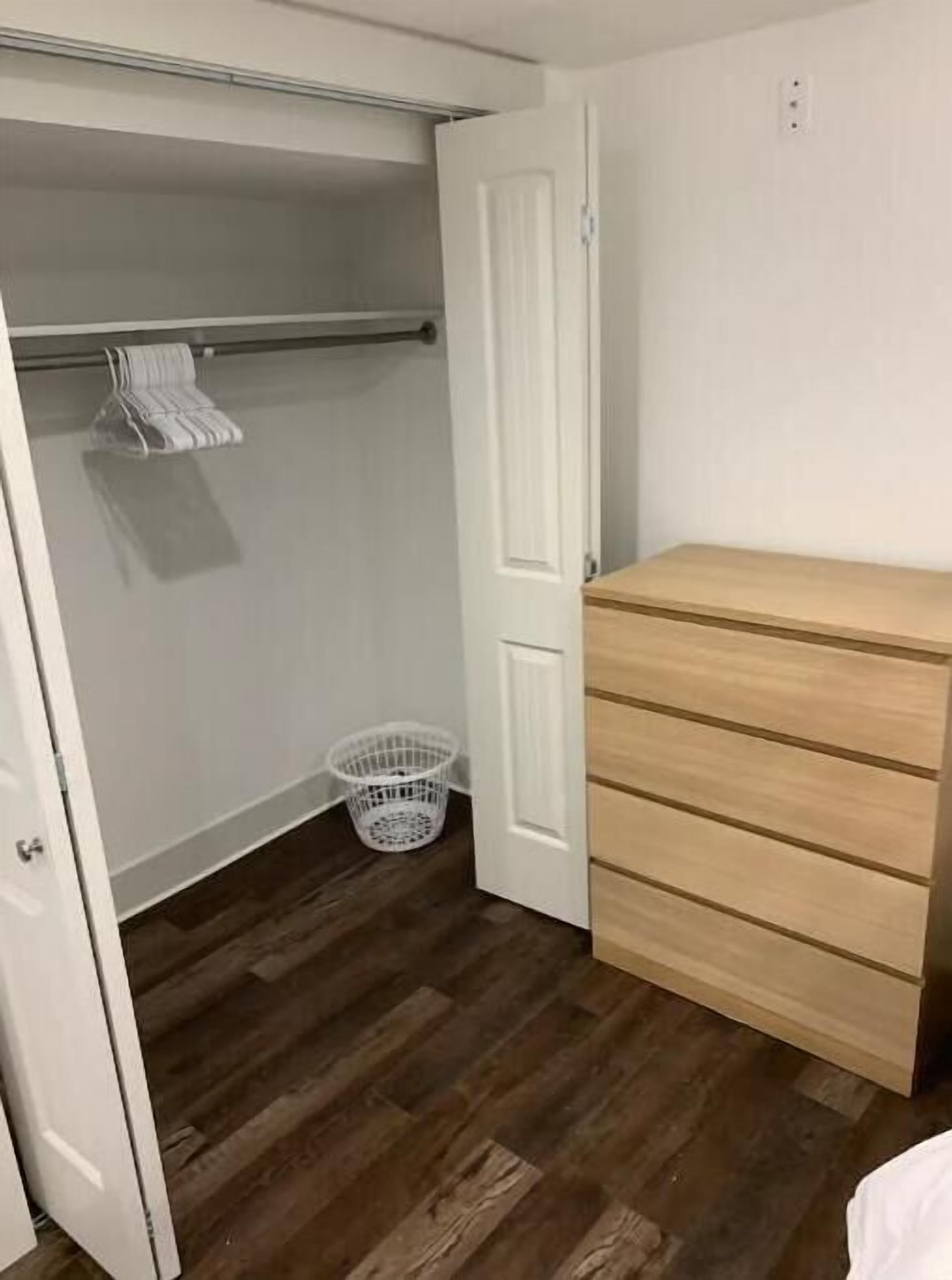 A bedroom with a wooden dresser, a closet with a hanging rod, and a laundry basket on the floor.