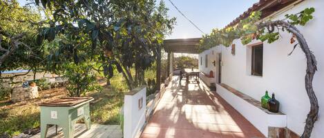 Un pasillo con piso de baldosas, un banco y una mesa a la izquierda. Hay plantas y una pérgola a la derecha.