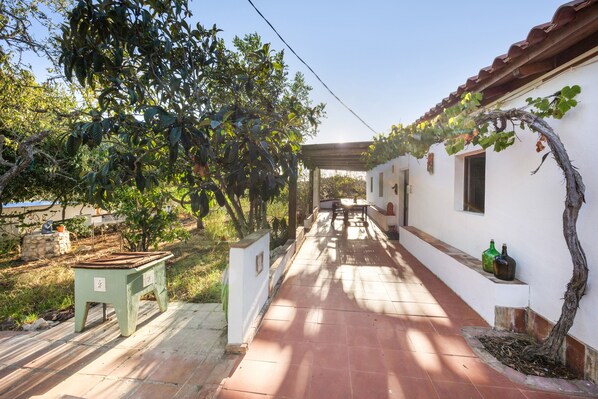 Un pasillo con piso de baldosas, un banco y una mesa a la izquierda. Hay plantas y una pérgola a la derecha.