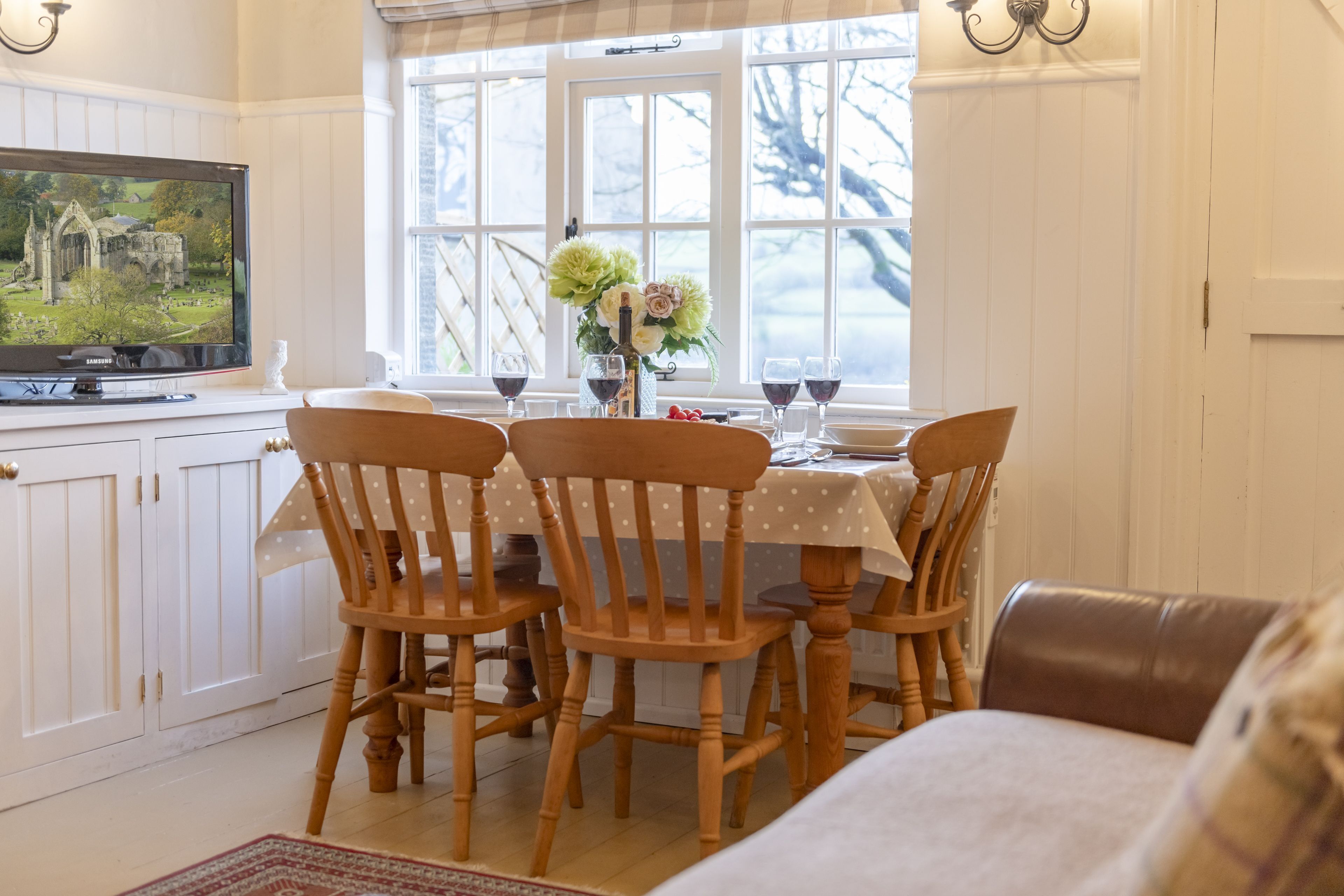 A dining area with a table set for four, wooden chairs, a television on a cabinet, and a window with a view of trees.