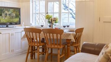 A dining area with a table set for four, wooden chairs, a television on a cabinet, and a window with a view of trees.