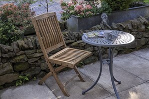 Ein Holzstuhl mit querverlaufenden Leisten an Sitz und Rückenlehne, neben einem runden Metalltisch mit einer Tasse und einer Zeitschrift.
