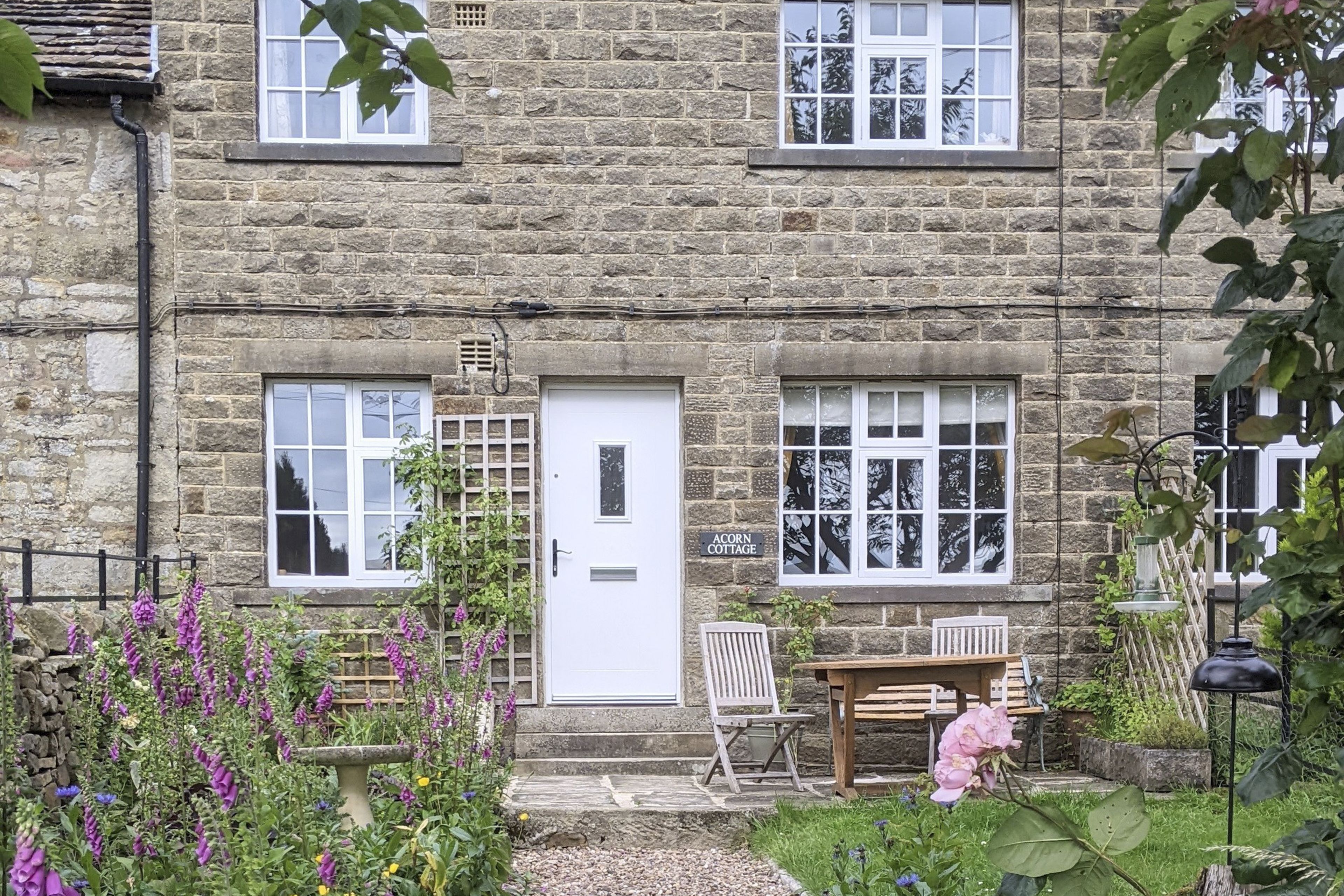 A stone cottage with a white door, two windows, and a small garden with purple flowers.