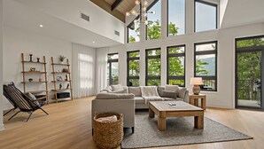 Ein geräumiges Wohnzimmer mit einer großen Couch, einem Couchtisch und Holzboden. Mehrere Fenster sorgen für natürliches Licht und Blick ins Grüne.