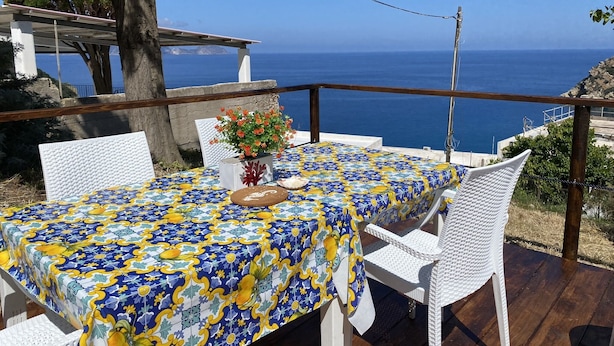 Une table dressée en plein air, avec vue sur la mer, des chaises en osier blanches et une nappe à motifs.