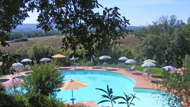 A pool with sun loungers and umbrellas, surrounded by greenery and hills in the background.
