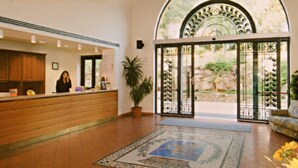A hotel reception area with a receptionist, a potted plant, and a large arched doorway leading outside.