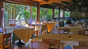 An outdoor restaurant with tables covered in beige tablecloths, wooden chairs, and a view of trees through the open sides.