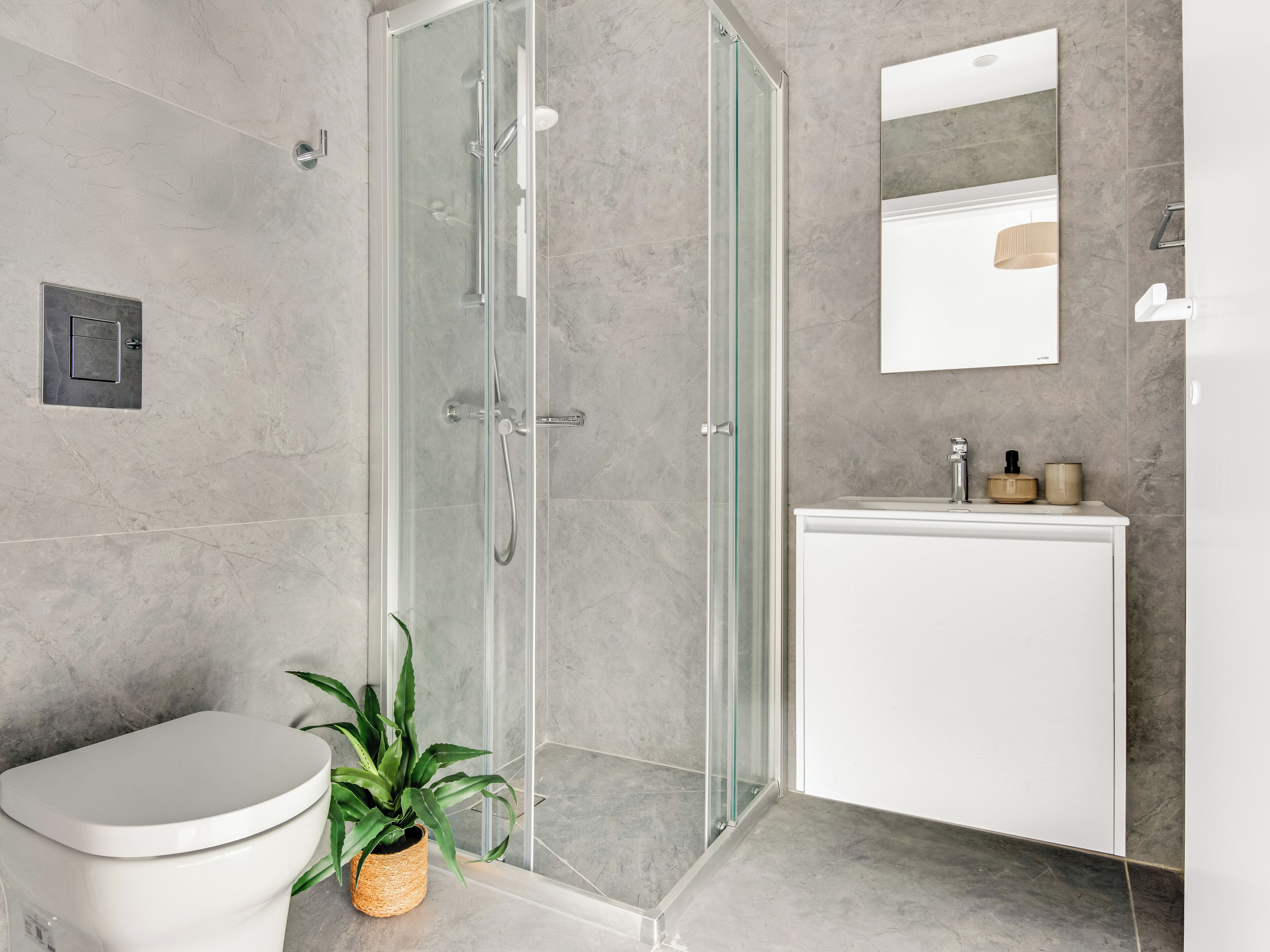 Un baño moderno con ducha envuelta en vidrio, inodoro blanco, lavamanos con espejo y una planta en maceta.