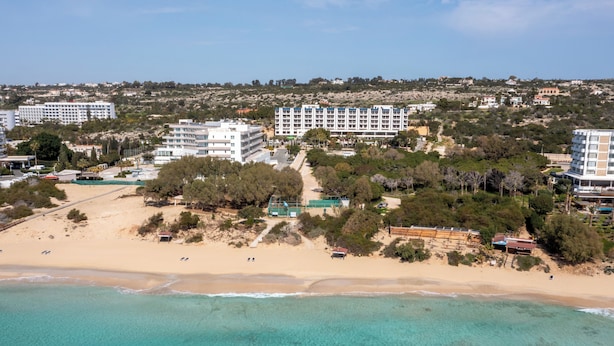 An aerial view of a coastal area with a beach, several buildings, and a clear blue sea. - Sanders Florida - Trendy 1-bdr Apt w Sea View (Ayia Napa)