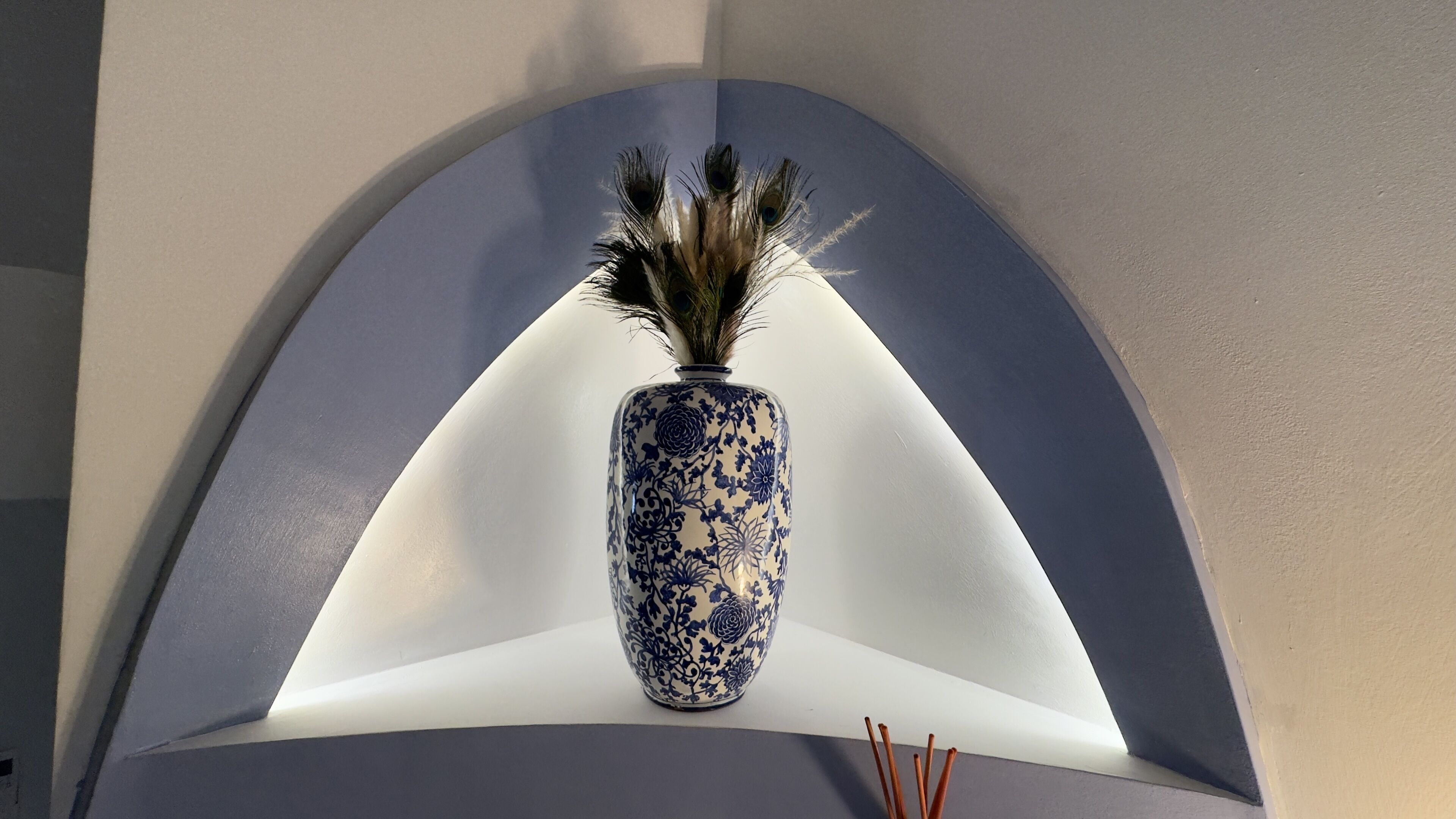 Un vase bleu et blanc orné de motifs floraux, surmonté d’une composition de plumes, le tout posé sur un piédestal blanc sous un plafond cintré.