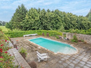 Una piscina rectangular con solárium de piedra, rodeada por un seto y un muro de piedra.