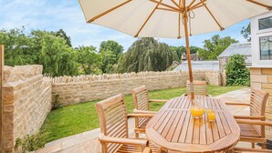 An outdoor wooden table and chairs with a large umbrella, surrounded by stone walls and greenery.