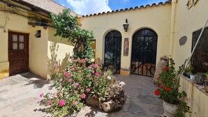 Un patio con un edificio amarillo, una puerta de madera y un portón negro. Hay plantas y flores en macetas.