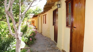 Un estrecho sendero de piedra flanqueado por edificios con puertas y ventanas de madera, rodeado de follaje y flores.