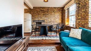 A cozy living space with a brick wall, a blue sofa, a round table with chairs, and a small kitchen area with a refrigerator. - Charming Shoreditch Apartment (London)