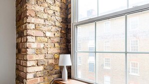 A room with a brick wall, a window with a view of buildings, and a white lamp on a shelf. - Charming Shoreditch Apartment (London)
